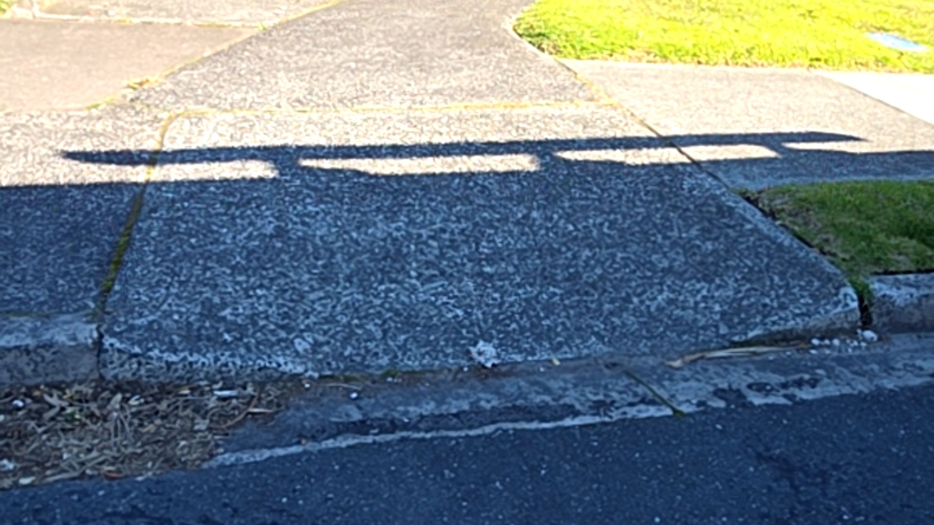 Wide grey aggregate concrete kerb ramp with a gentle gradient connecting a footpath to an asphalt road. A long horizontal shadow crosses the textured surface.