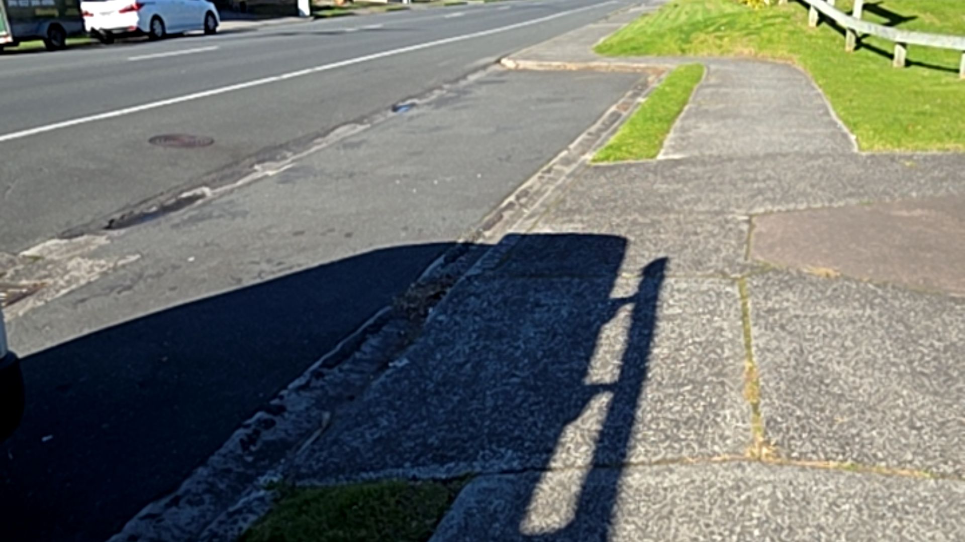 Wide, level concrete footpath with a coarse surface and expansion joints. It runs parallel to an asphalt road and is bordered by a grass verge and wooden post-and-rail fence.