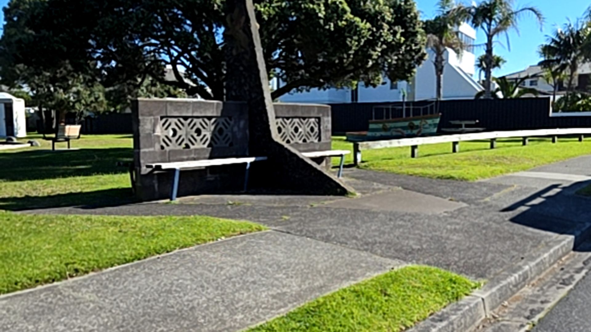 Level concrete pathway leads to two white benches with blue legs, set against a dark grey stone wall with cutouts. The area is surrounded by grass and a low white wooden fence.