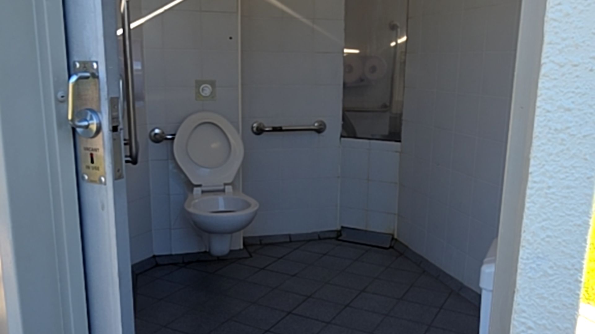 Level grey tiled floor leads to a wall-hung toilet with horizontal grab bars on both sides and a circular flush button. Wide door has a metal lever handle and vacancy indicator.