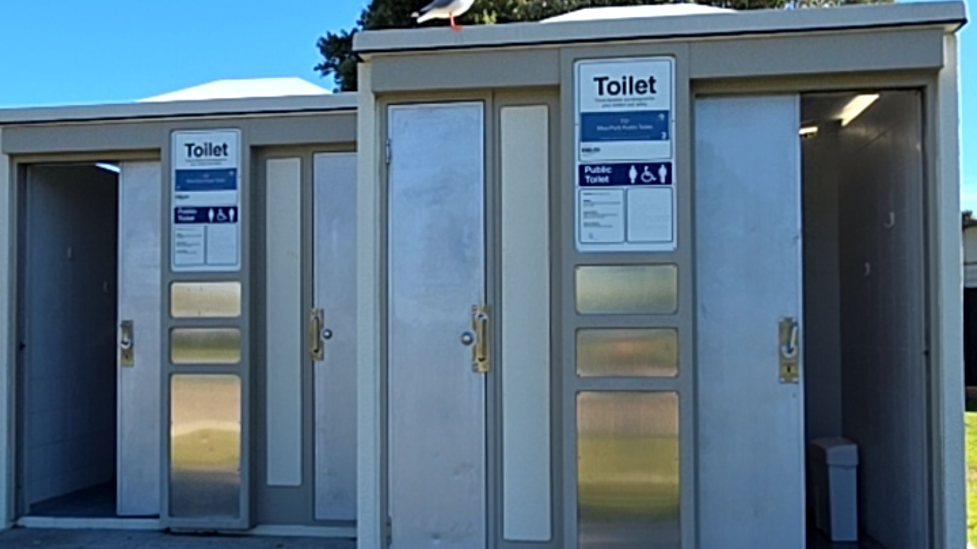 Level concrete path leads to light grey public toilet cubicles with wheelchair symbols. Silver metal sliding doors have vertical grab handles. Entry to the interior floor is level.