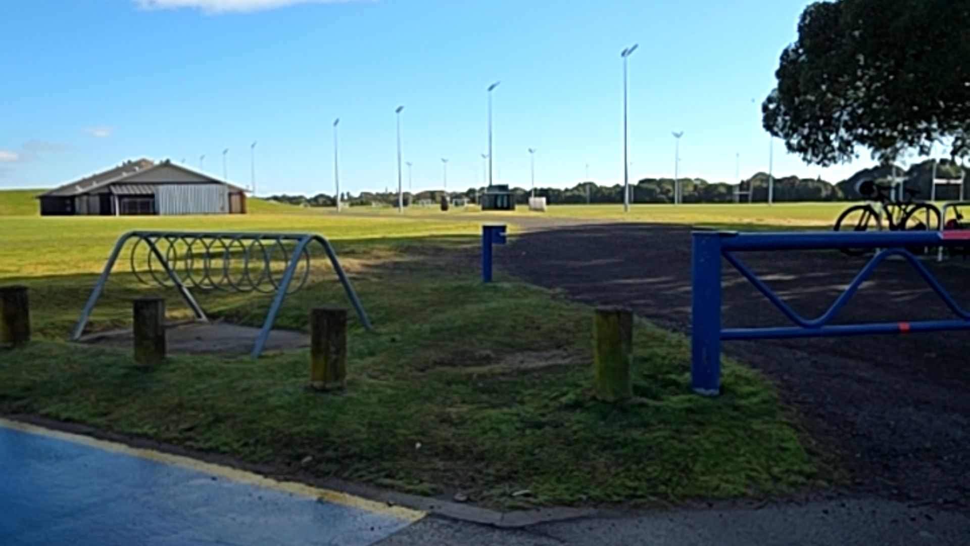 Level unsealed path blocked by a blue metal gate. Metal bike rack on grass behind wooden bollards. Wide, flat green fields lead to a grey pavilion. Wet asphalt surface with yellow line edge.
