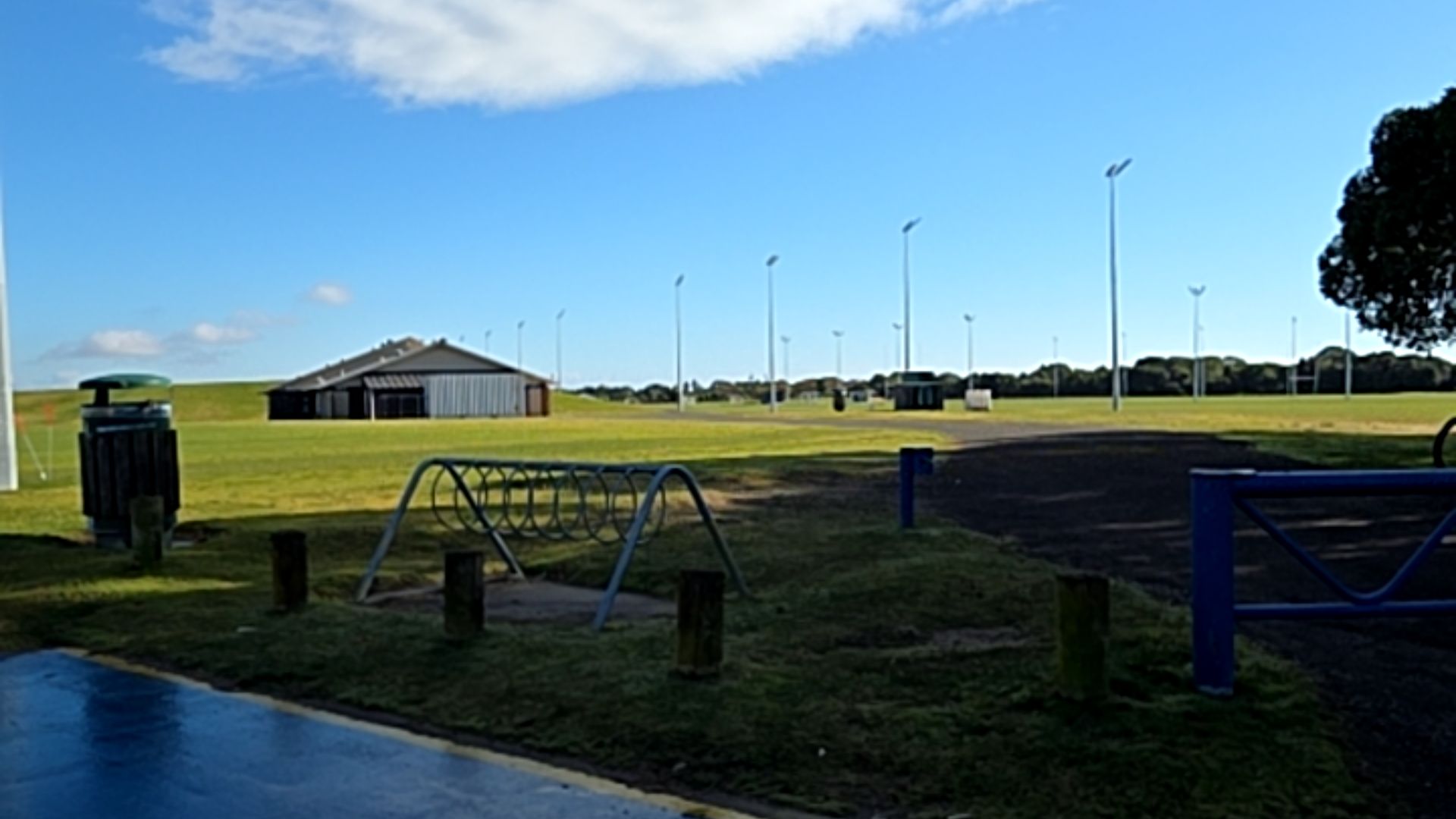 Bitumen path meets blue surface with yellow line. Grey metal bike rack and wooden bollards sit on grass nearby. Blue metal gate on right. Grassy field with tall floodlights and building beyond.