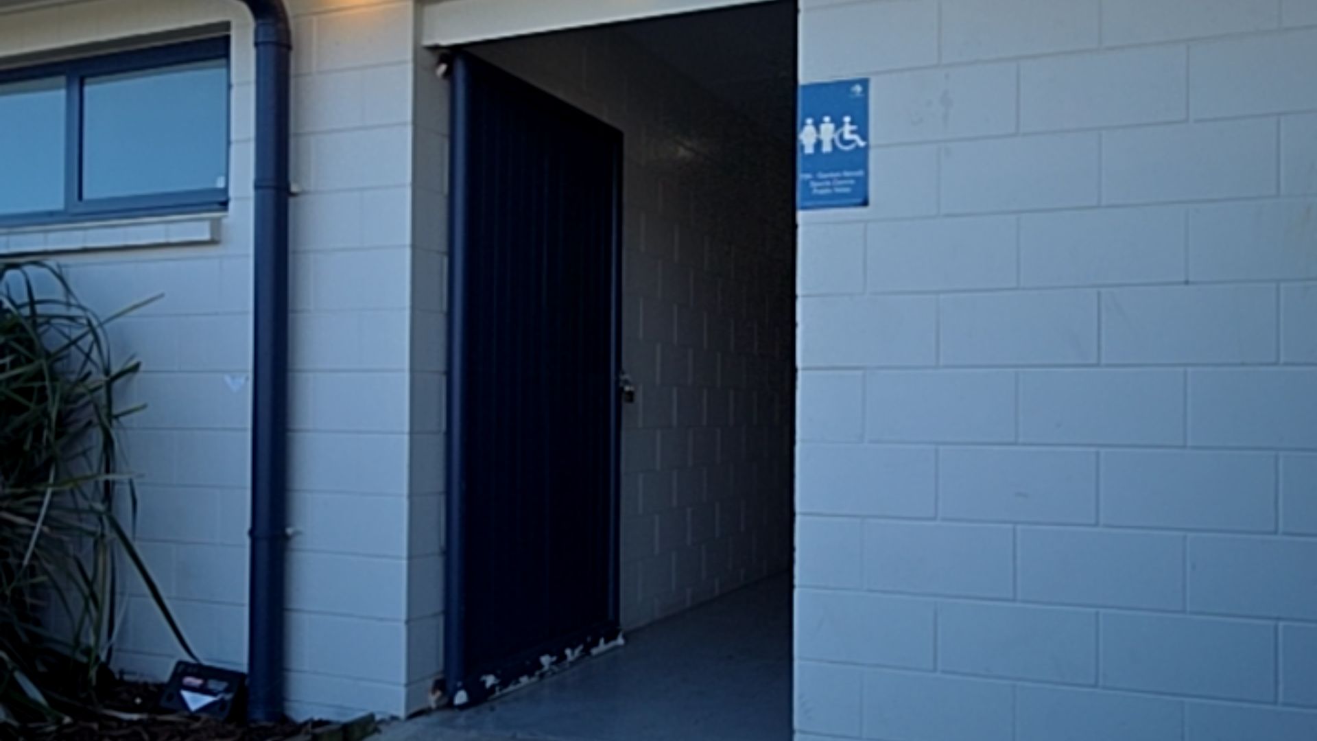 Level concrete entrance with a dark blue sliding door. White block-work walls feature a blue sign with white male, female, and wheelchair accessibility symbols.