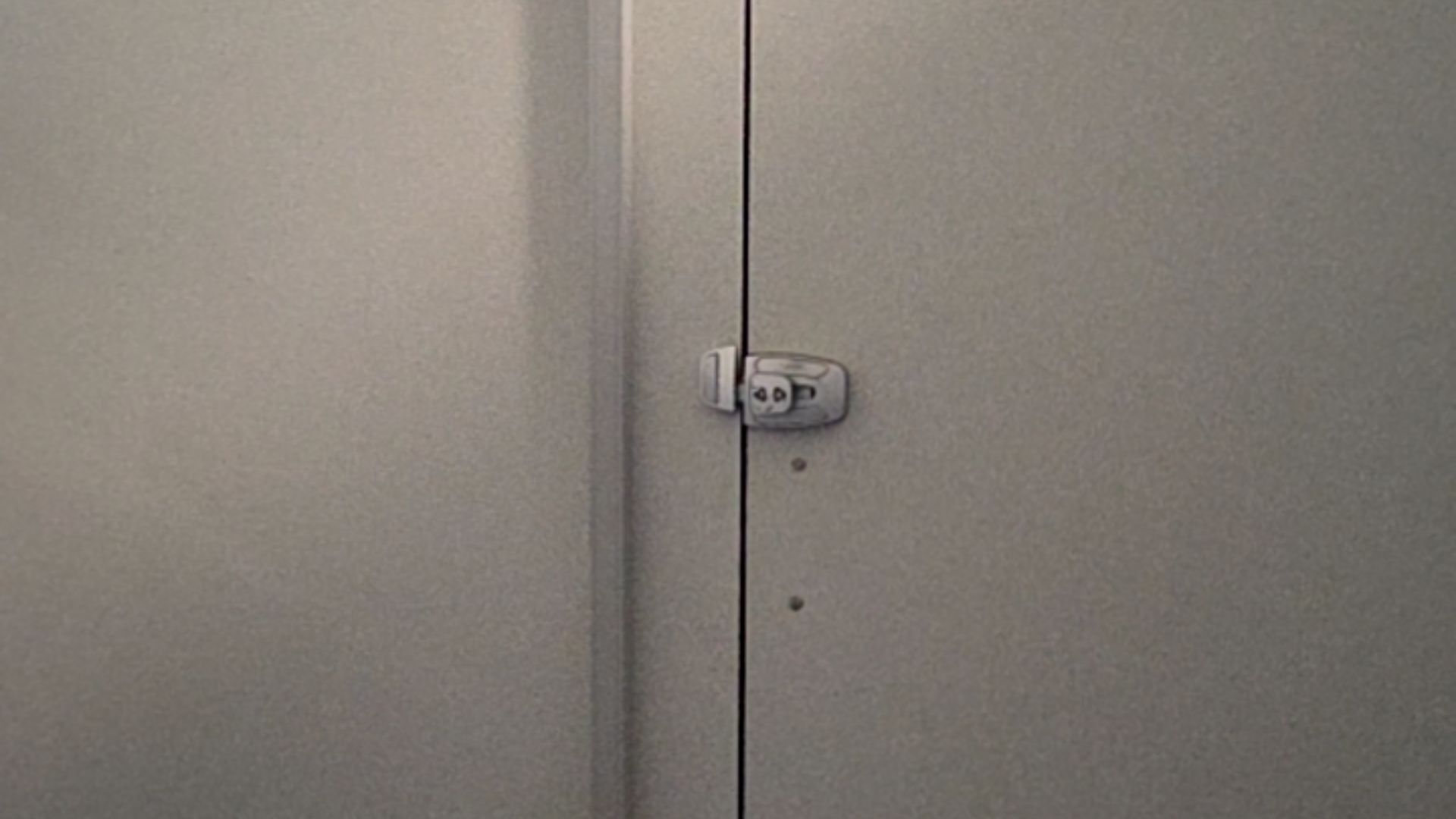 Grey toilet cubicle door with a silver metal slide latch. The latch is engaged across a narrow vertical gap between the door and the partition.