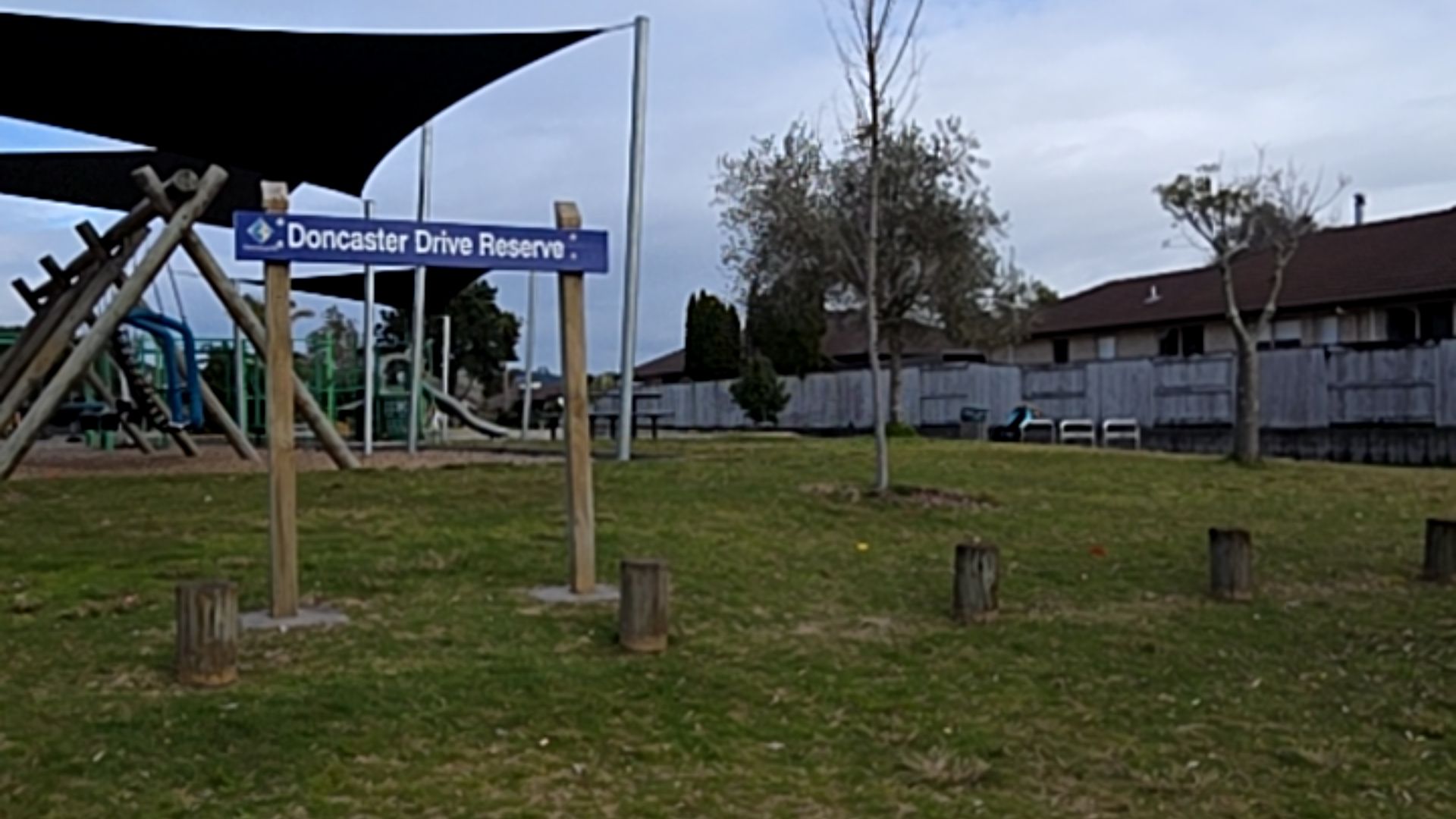 Doncaster Drive Reserve sign on timber posts. Grassy area with low wooden bollards. Background includes a playground with black shade sails, park benches, and a grey timber fence.