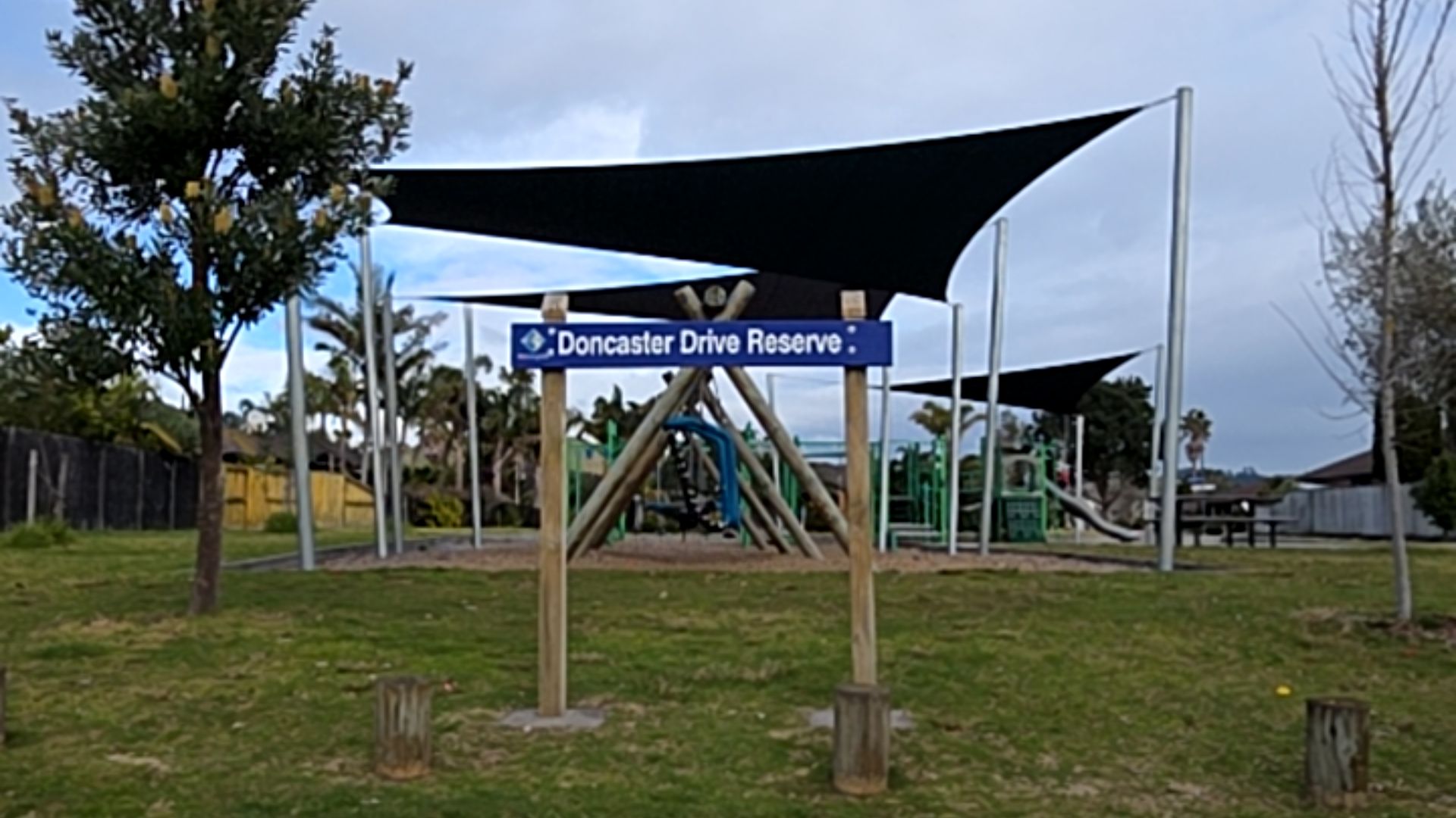 Blue 'Doncaster Drive Reserve' sign on wooden posts over grass. Behind, black shade sails cover play equipment on a bark surface. Three wooden stumps are in the grass. No paved paths visible.