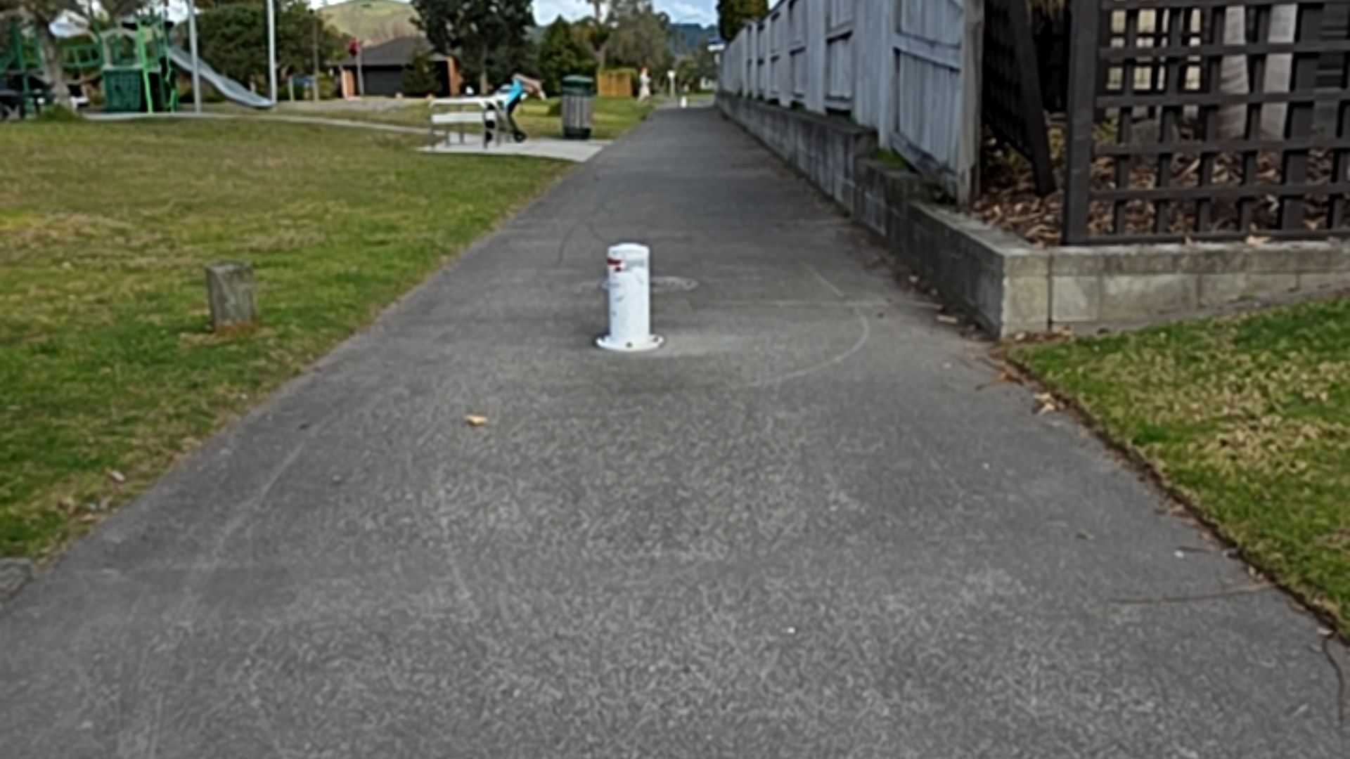 Wide level concrete pathway with a white bollard in the centre. Grassy area and playground to the left, and a timber fence on the right.