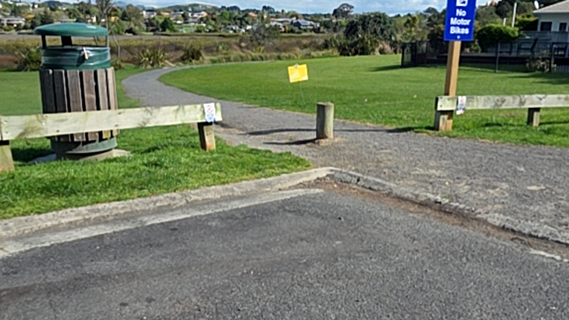 Level gravel path meets an asphalt surface, narrowed by a central wooden bollard and side fences. A green rubbish bin is on the left and a blue "No Motor Bikes" sign is on the right.