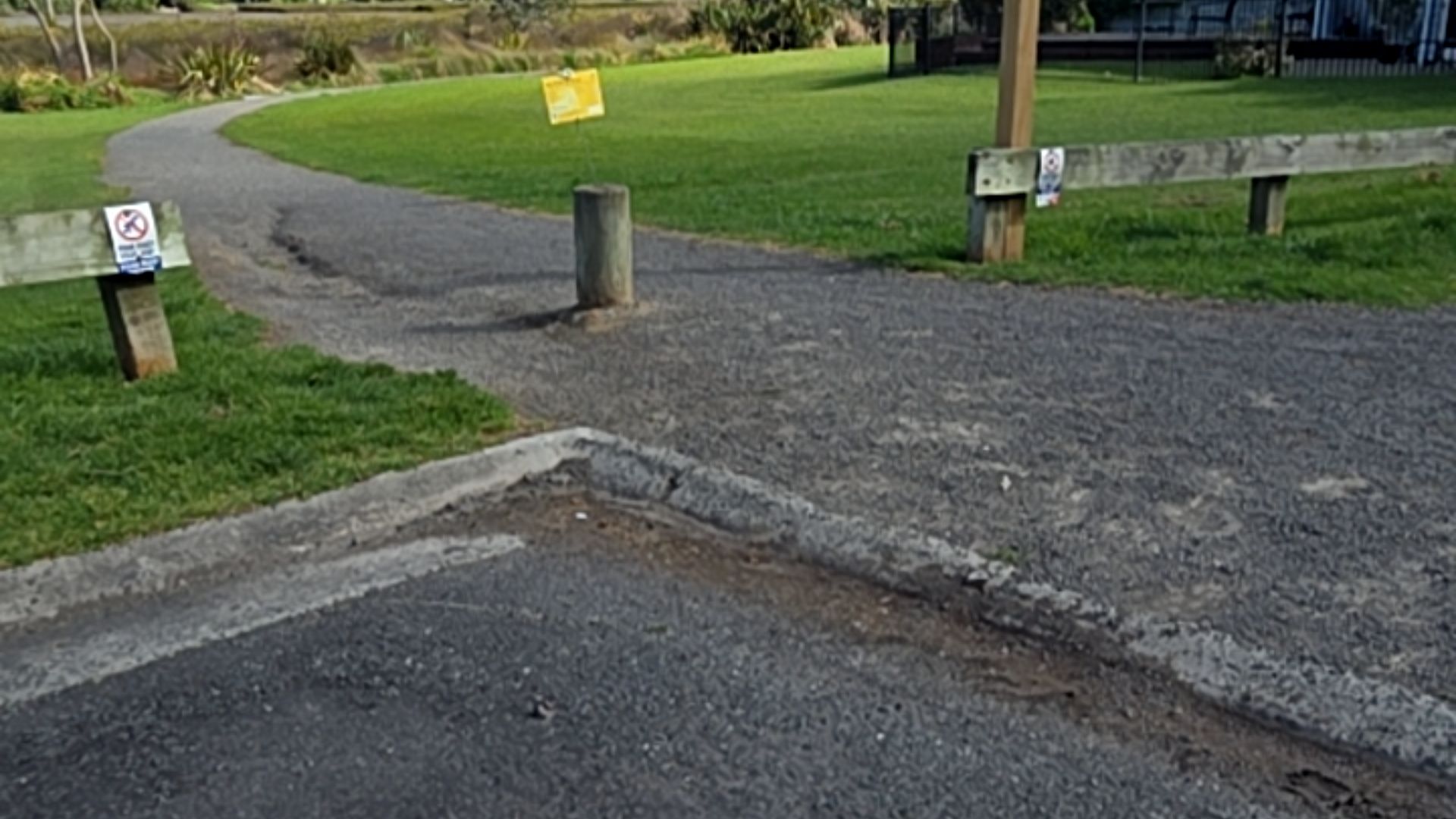 Level asphalt area leads over a concrete kerb to a wide grey gravel path. A single wooden bollard stands in the centre of the path entrance between low timber fences.