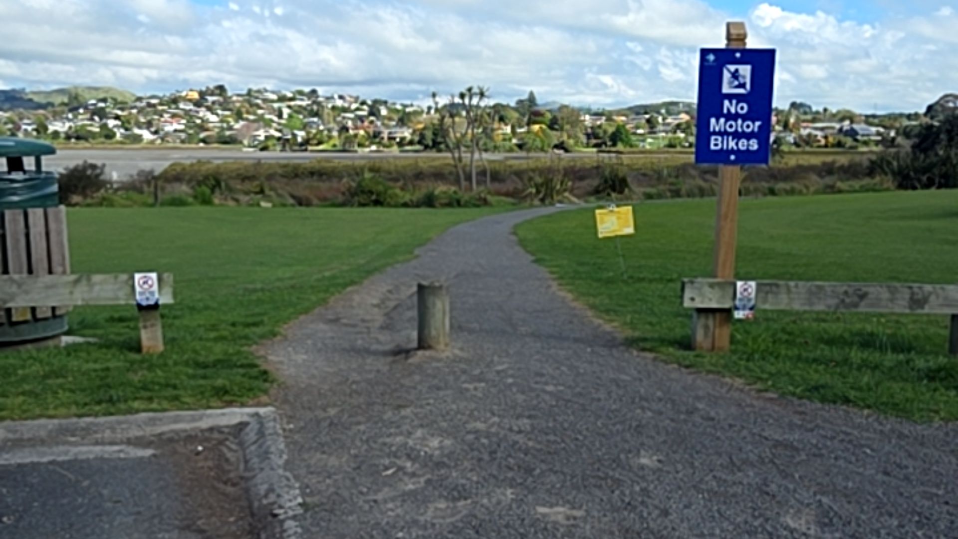 Level gravel path with a central grey bollard at the entrance between low timber fences. A blue "No Motor Bikes" sign stands on the right. Green grass borders the path.