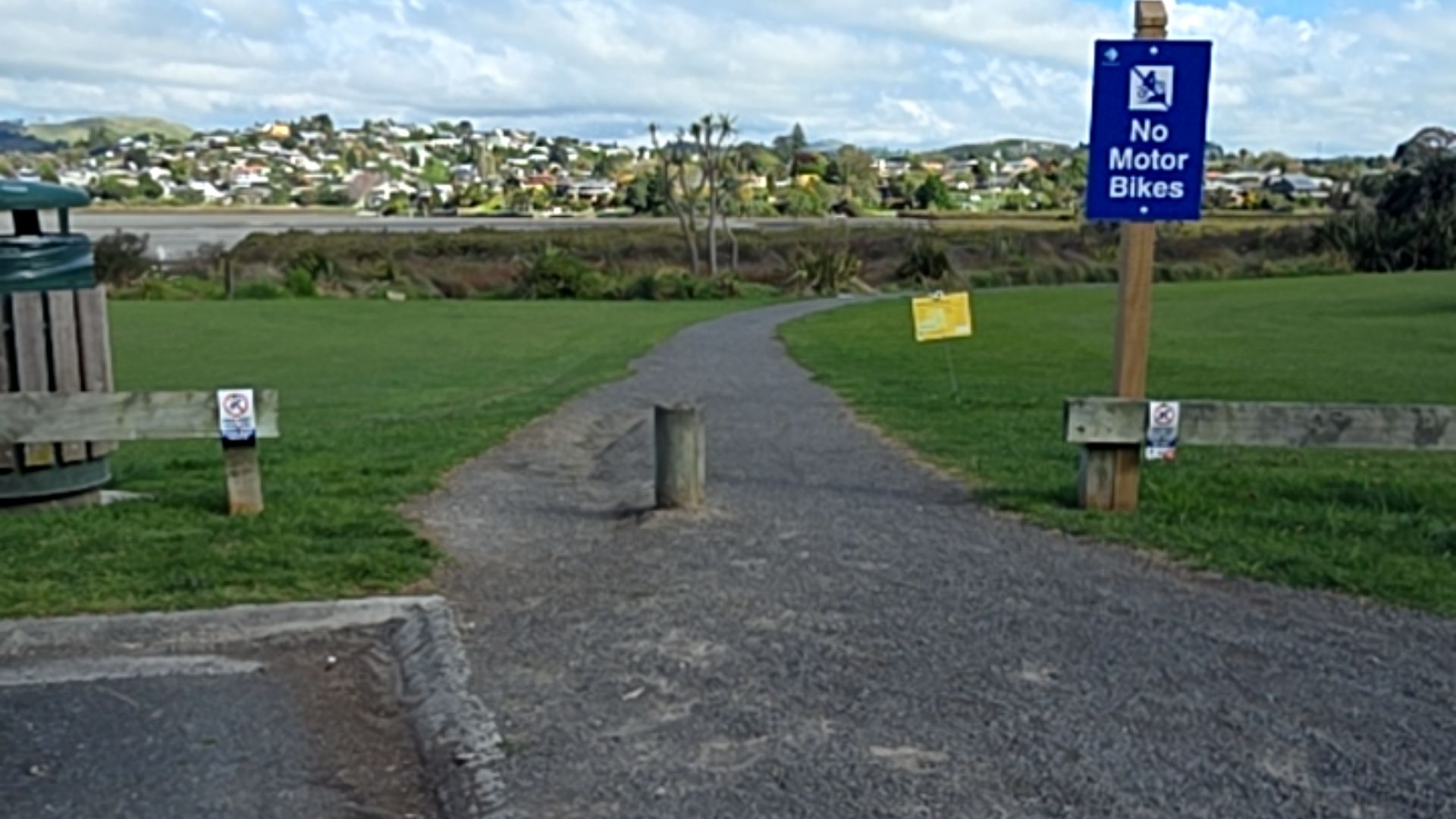 Wide, level gravel path with a central wooden bollard. Flanked by low timber fences. A tall wooden post to the right displays a blue 'No Motor Bikes' sign.