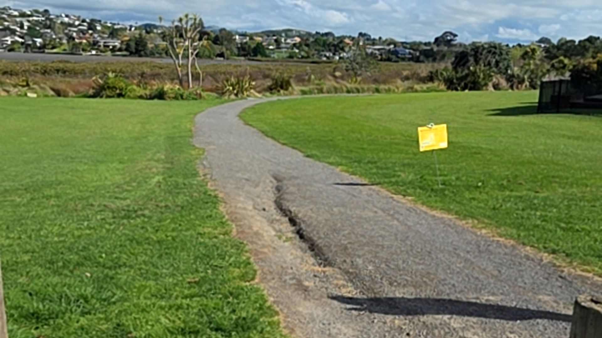 Uneven grey gravel pathway through green grass. A central rut is visible on the surface. A small yellow sign on a post stands to the right of the path.