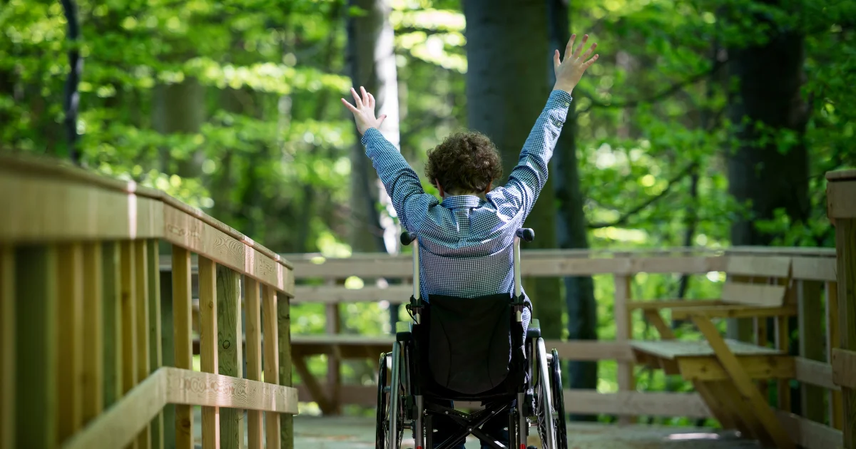 A young boy in a wheelchair on a wooden walkway surrounded by trees.