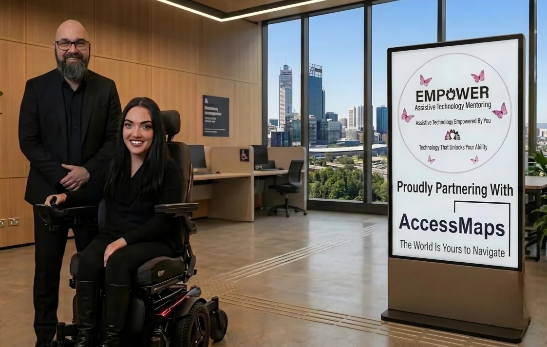 A man and a woman smile in a modern office setting, standing next to a large digital display panel. The man is bald with a dark beard and glasses, dressed in a black suit. The woman has long dark hair, is wearing black clothing, and is using a motorized wheelchair. They are positioned in the foreground on the left. The background features light wood wall panelling, office workstations, and large windows revealing a sunny Perth city skyline. The vertical digital display panel on the right features a white screen with two logos. The top logo is a circle surrounded by pink butterflies, containing the text EMPOWER Assistive Technology Mentoring, followed by the taglines Assistive Technology Empowered By You and Technology That Unlocks Your Ability. Below this, the display reads Proudly Partnering With, followed by the AccessMaps logo and the tagline The World Is Yours to Navigate.