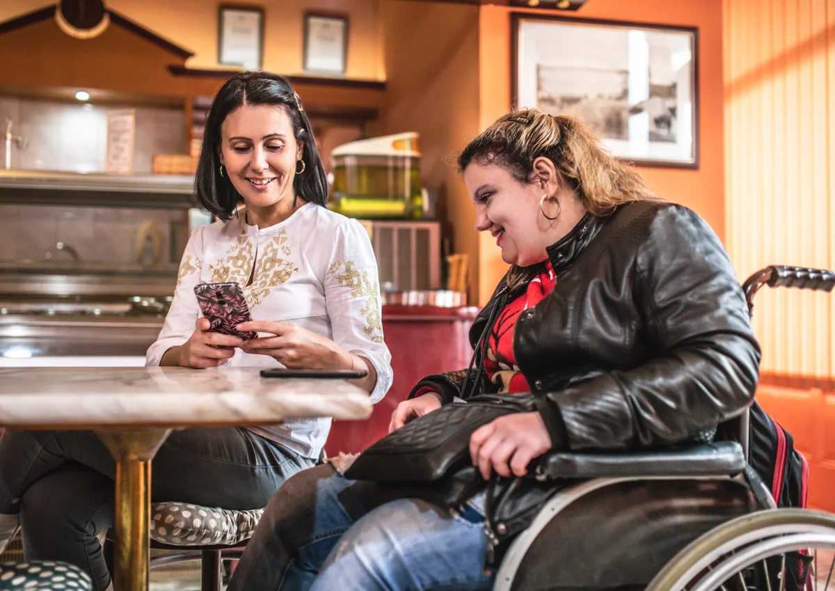 Two people sitting at a table. One in a wheelchair and one sitting on a chair. They were both looking at a smartphone, looking through their options.