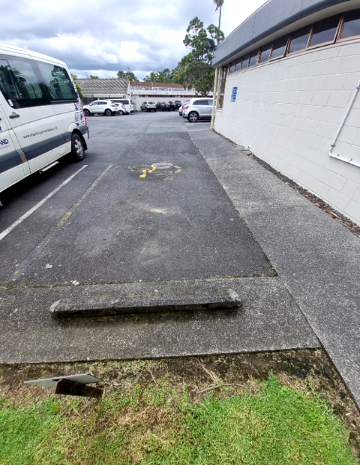Level asphalt accessible parking space with a yellow wheelchair symbol and concrete wheel stop. A concrete pathway runs beside the space, adjacent to a light-coloured brick building.