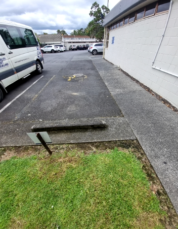 Asphalt parking bay marked with a yellow wheelchair symbol. A level concrete pathway runs beside a white block building. A concrete wheel stop sits at the end near a grassed area.