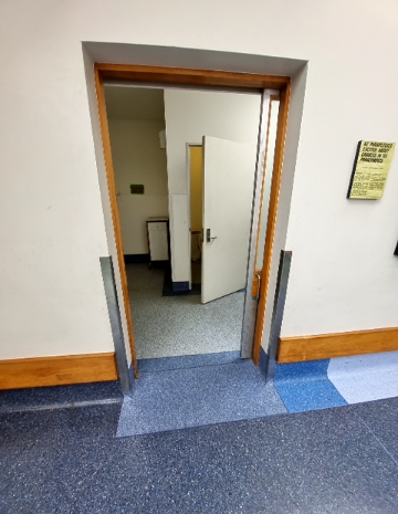 Wide timber-framed doorway with blue ramped floor surface and stainless steel corner guards. Interior features grey speckled flooring, a white cabinet, and another open white door.