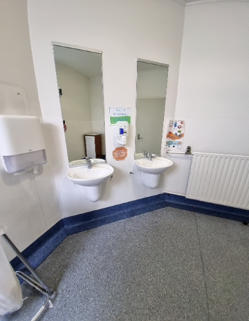 Two white wall-mounted basins at staggered heights. Level grey speckled floor. Soap and paper towel dispensers. 'Hand Washing Only' sign between tall mirrors. White radiator on the right.
