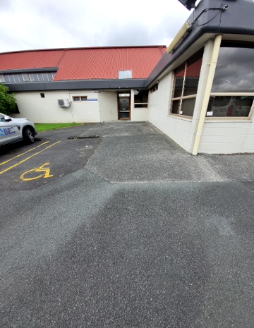 Accessible parking space with yellow wheelchair symbol on dark asphalt. A wide, level concrete pathway leads to a single glass door entrance of a cream-coloured building with a red roof.