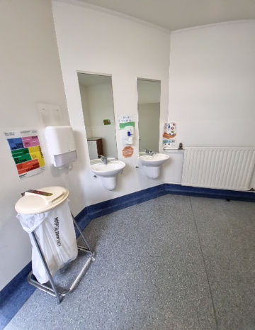 Wall-mounted white basins with lever taps and mirrors. Level grey speckled non-slip floor. Metal grab rail beside one basin. White walls feature signage, dispensers, and a mobile waste bin.