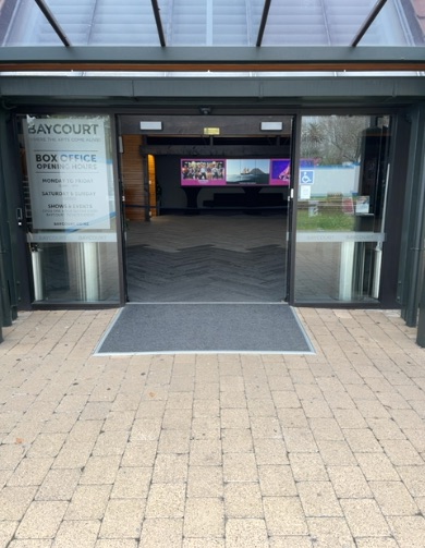 Wide, level automatic glass doors open into Baycourt. The pathway outside is light-coloured paved, continuing to a dark mat inside. A wheelchair access symbol is visible on the right door.