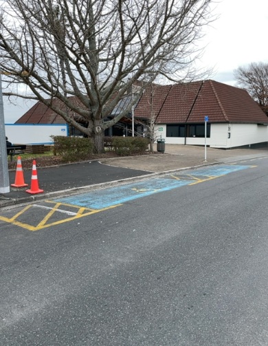 Blue-marked disabled parking bay on an asphalt road with a pole sign. Two orange cones obstruct the concrete kerb nearby, in front of a building with a brown tiled roof.