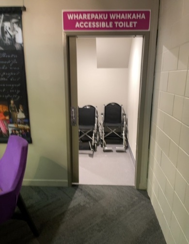 Pink 'ACCESSIBLE TOILET' sign above an open doorway. Dark grey carpet transitions to light grey flooring inside, where two folded wheelchairs are visible.