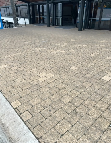 Multiple dark-framed glass doors lead to a modern building, fronted by a wide, level pathway of light brown pavers. A concrete kerb is on the left.