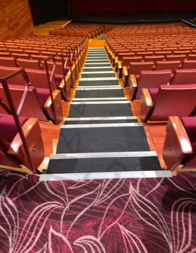 Wide central staircase with dark grey steps and white non-slip strips, flanked by tiered red upholstered seating. Wooden balustrades line the lower steps. Burgundy patterned carpet in foreground.