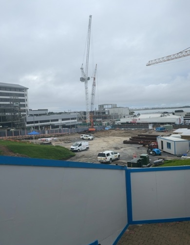 Active construction site with uneven dirt ground, featuring multiple vehicles, portable buildings, and tall cranes. A foreground fence partially obscures a brick pathway and grassy mounds.