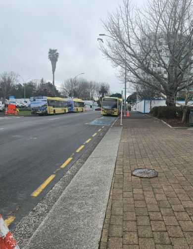 Level, brown paved footpath and concrete strip next to a road with yellow lines. A blue marked area on the road suggests accessible parking or a bus stop zone near a damaged yellow bus. Cones are p...