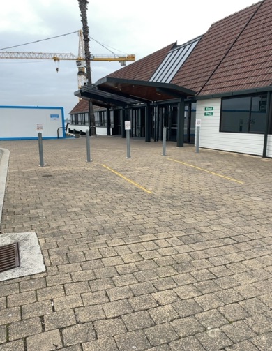 Level paved forecourt with grey bollards leading to wide glass entrance doors under a dark canopy. Green signs are visible on the building to the right.