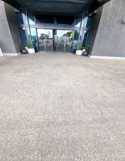 Wide, level grey concrete pathway leading to a modern building with automatic glass sliding doors. Potted plants flank the entrance.