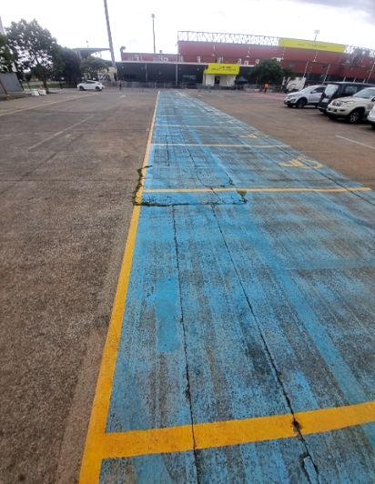 Worn, blue-painted concrete pathway with yellow markings in a car park. Minor cracks are visible on its surface. It leads towards a building and parked cars in the distance.