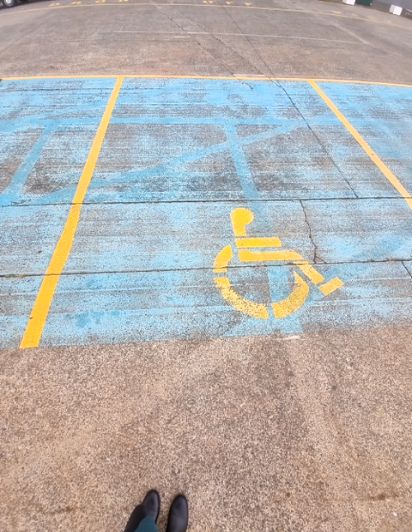 Accessible concrete parking bay with faded blue paint and yellow wheelchair symbol. Yellow lines mark the space, and the surface shows cracks.