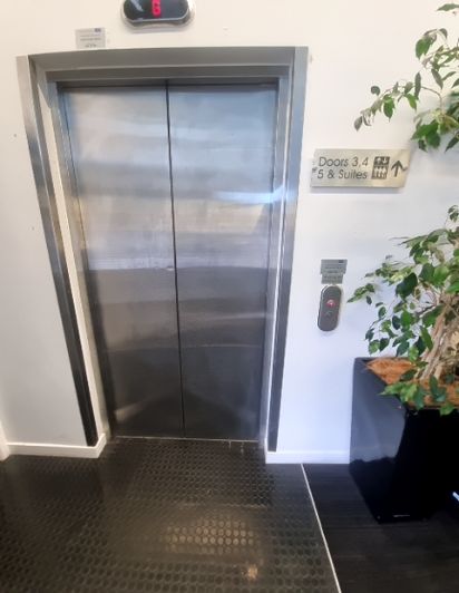 Stainless steel lift on a level, dark hexagonal tiled floor. Red digital display shows '6'. Illuminated red 'up' call button on right. Signage directs to 'Doors 3,4' & '5 & Suites' upstairs.