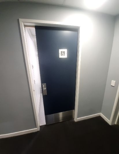 Dark blue accessible door, slightly ajar, with a wheelchair symbol, silver kick plate, and handle in a grey hallway with a dark floor.