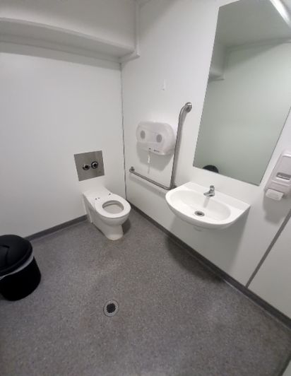 White toilet room with a wall-mounted toilet & basin. Includes two chrome grab bars for support and a level grey non-slip floor with a drain.
