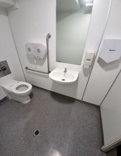 Accessible toilet with dark grey non-slip floor and white walls. Includes toilet, basin, mirror, paper towel dispenser, Dyson hand dryer, and stainless steel grab rails by toilet and basin.