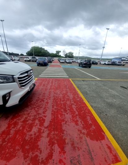 Worn, red pathway with yellow border lines, possibly wet, leads to a zebra crossing within a large outdoor car park. Many vehicles are parked under a cloudy sky.