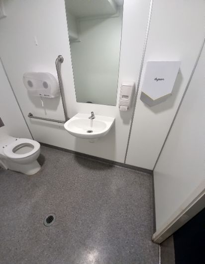 Accessible toilet with grab bars by toilet and wall-mounted basin. White walls, grey non-slip floor with drain, paper towel dispenser, soap dispenser, and Dyson hand dryer.
