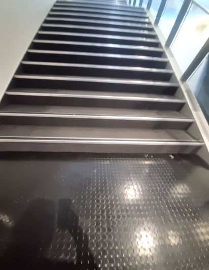 Steep, dark grey staircase with silver non-slip strips on each tread. At the bottom, a dark grey rubber floor with a studded texture for grip.