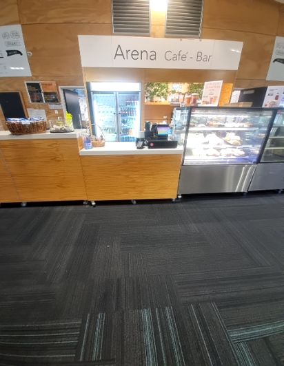 Arena Cafe-Bar counter, light timber-look, with glass food display. Dark grey patterned carpet floor, appearing level.