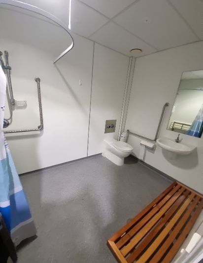 Accessible bathroom with a roll-in shower area, grab bars at the shower, toilet, and basin. Features a level grey floor and a wooden slatted bench.