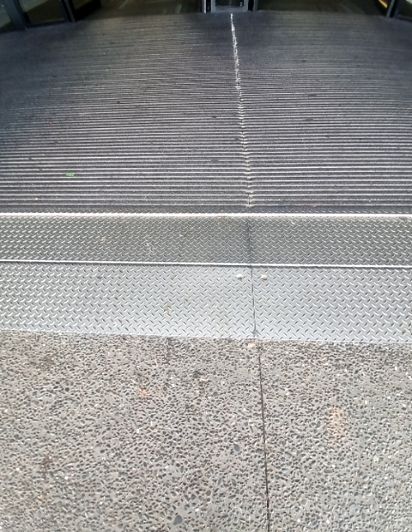 Level pathway transition: grey exposed aggregate concrete to silver diamond plate, then to wide grey corrugated matting leading to an entrance.
