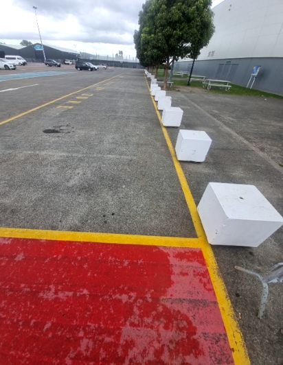 Level grey paved path with yellow markings and a red painted section. A line of white concrete bollards forms a barrier along the right edge. Cars and building in background.