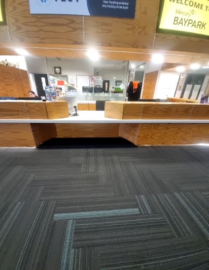 Level dark grey carpet-tiled floor with blue stripes leading to a light reception desk with wooden front panels and knee space underneath. Bright reception area, sign reads 'WELCOME TO BAYPARK'.