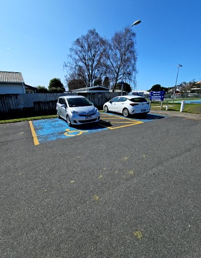 Two accessible parking spaces painted blue with yellow wheelchair symbols on level bitumen. A yellow-lined shared zone separates two white cars. Signage and a timber fence are in the background.